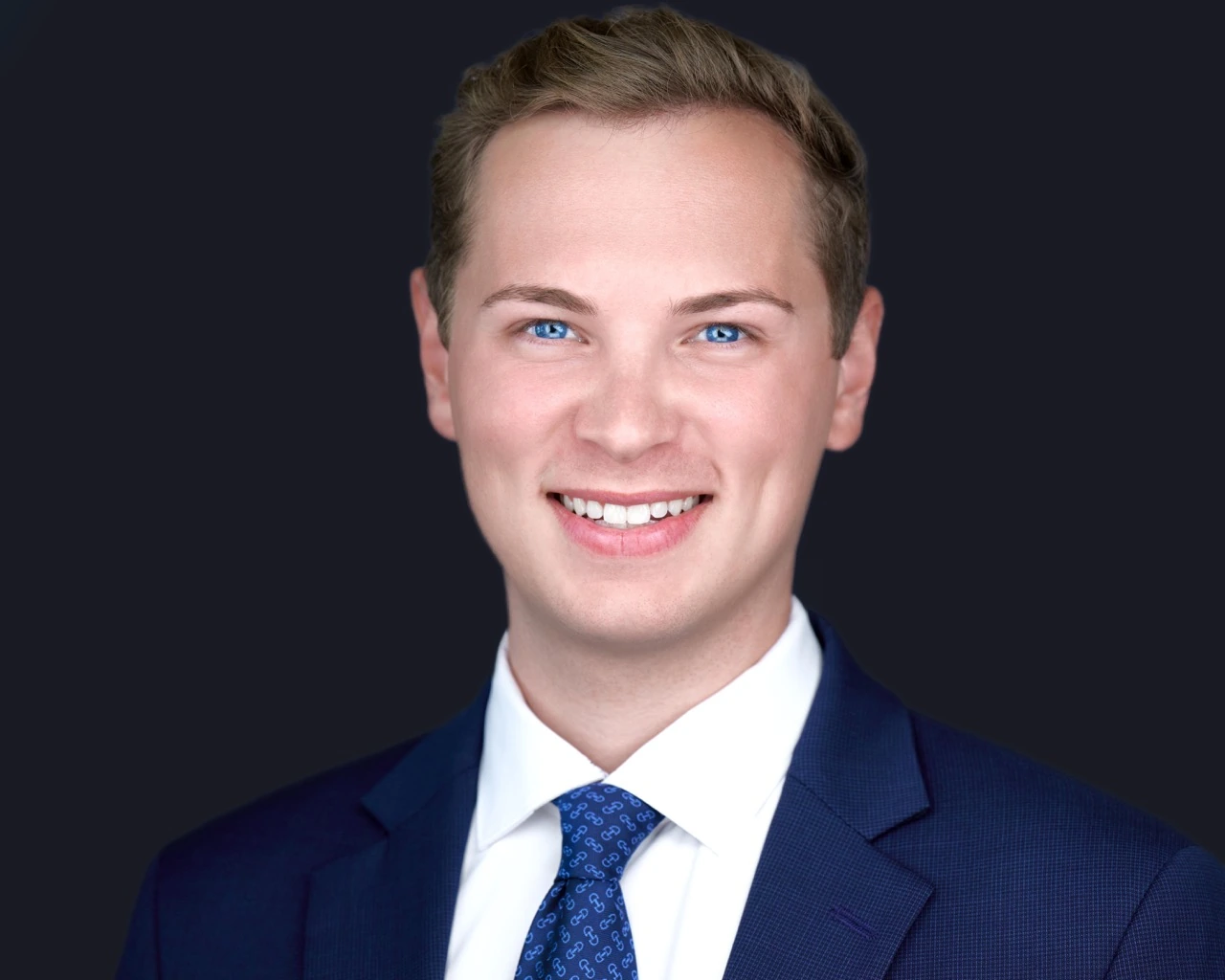 Professional male headshot by Deborah Heinlen Photography, Cincinnati — smiling young man in navy suit and blue patterned tie against dark studio background