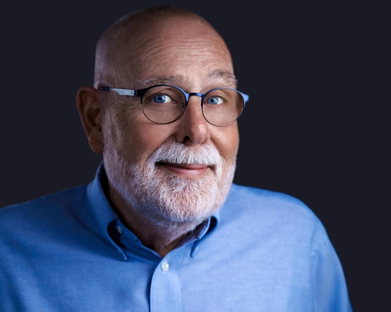 Professional male headshot by Deborah Heinlen Photography, Cincinnati — senior professional man with white beard and wire-rimmed glasses in light blue shirt against dark studio background