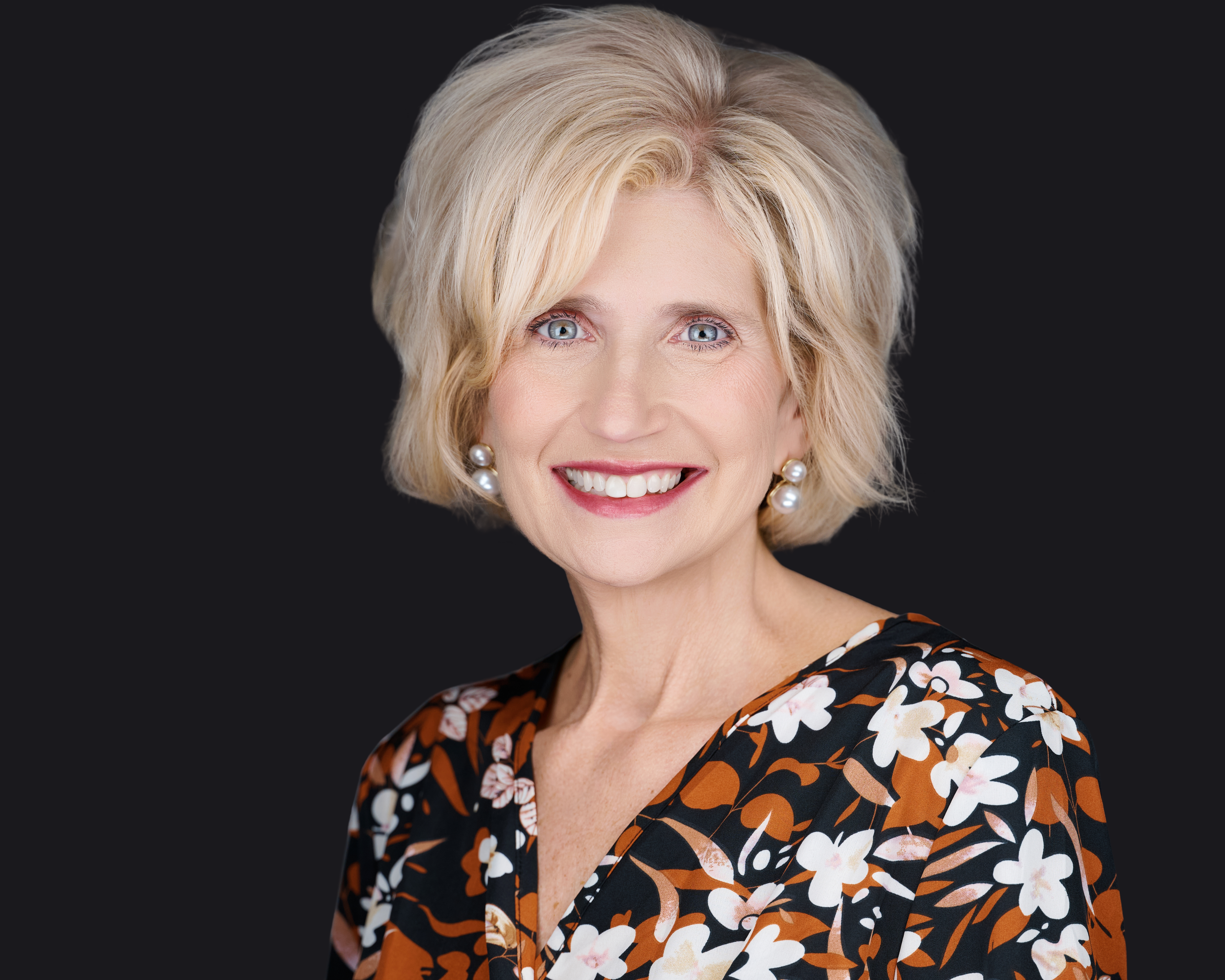 Professional female headshot by Deborah Heinlen Photography, Cincinnati, smiling woman with blonde hair in a floral top against dark studio background