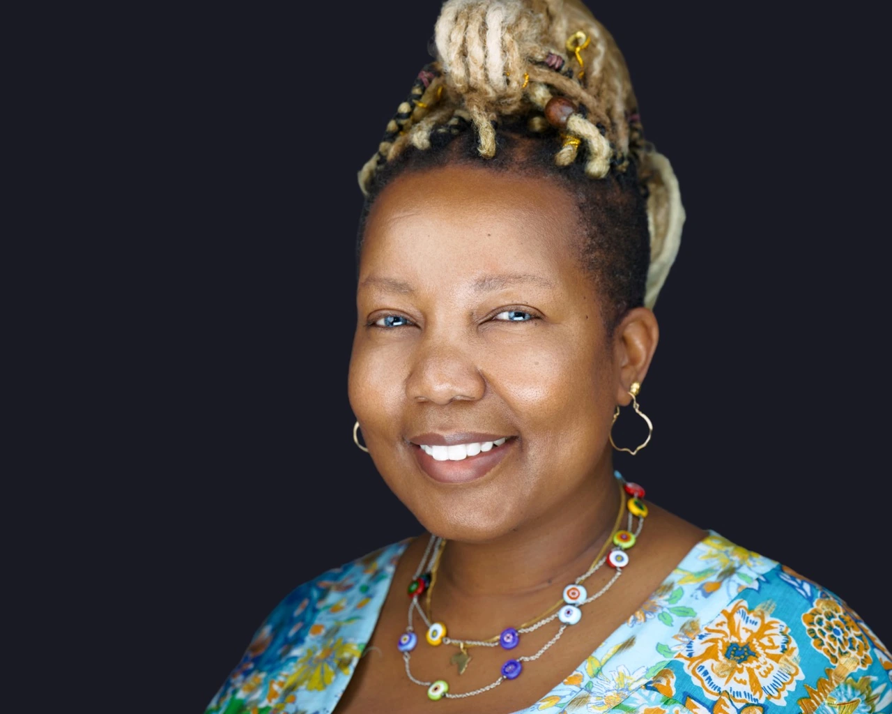 Professional female headshot by Deborah Heinlen Photography, Cincinnati — smiling woman with locs updo and colorful floral top and beaded jewelry against dark studio background