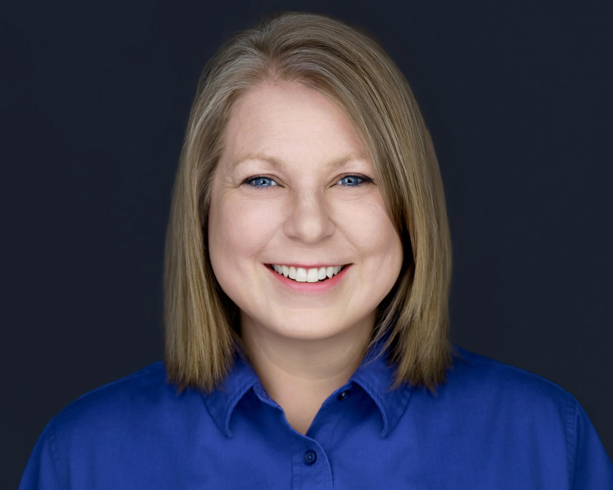 Professional female headshot by Deborah Heinlen Photography, Cincinnati — smiling woman with short blonde hair in blue shirt against dark studio background